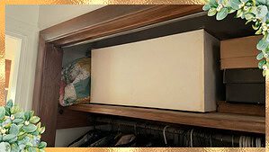 An old yellowed wedding dress preservation box sitting on a crowded hall closet shelf between a folded quilt and shoeboxes