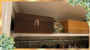 A worn wooden inherited silverware chest with teal felt lining sitting closed on a crowded hall closet shelf
