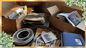 Old cardboard boxes and photo albums full of vintage family photographs stacked on the floor, with loose faded prints and a slide tray beside them