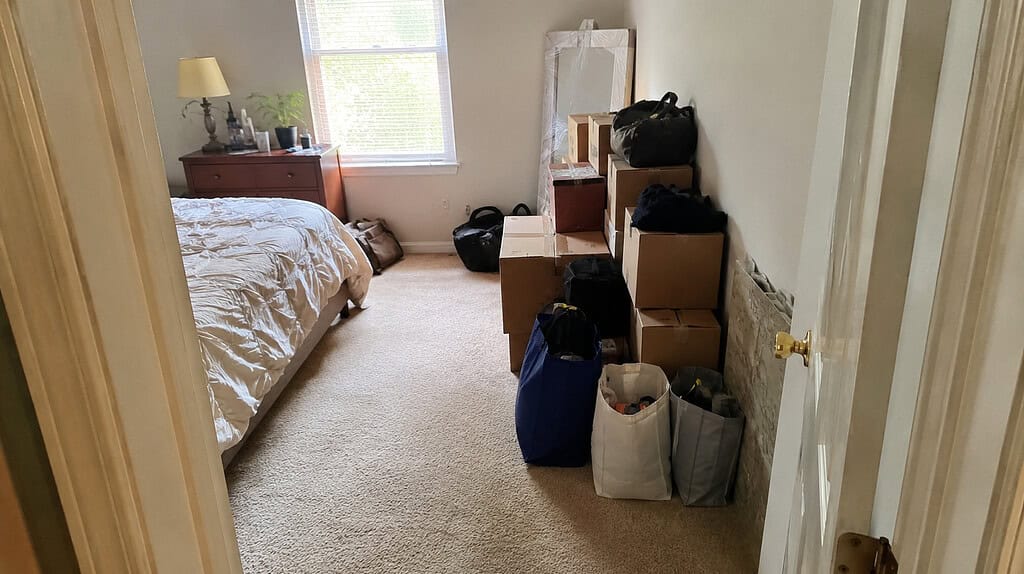A bedroom doorway looking into a room starting to fill up with boxes and bags along one wall, still walkable but getting crowded