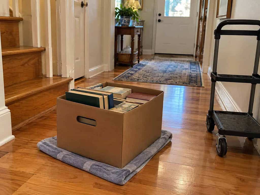 A box of books resting on an old towel on a wooden floor, ready to slide, with a small rolling cart nearby