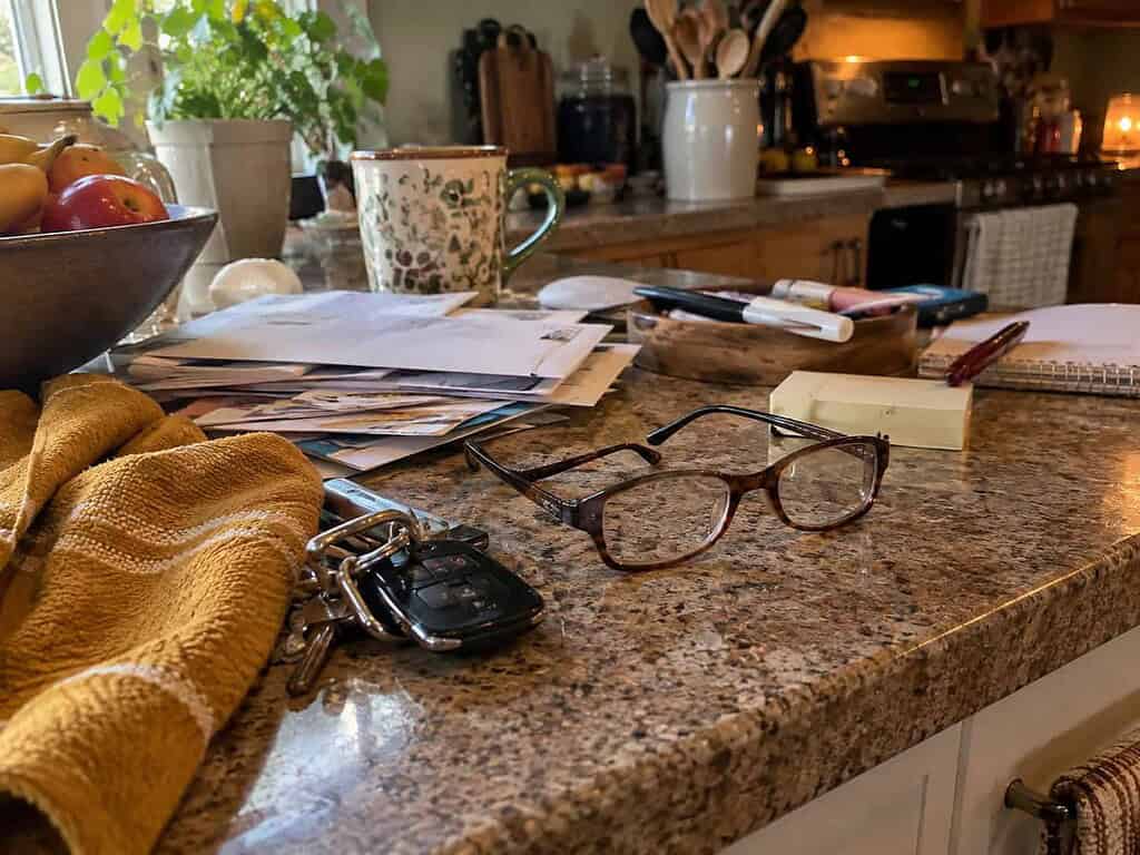 A cluttered kitchen counter with reading glasses set down among scattered mail and keys in an ordinary home