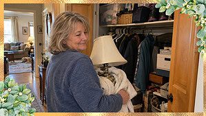 A woman in her 60s smiling softly at an overstuffed hall closet, holding an old lamp from her first apartment