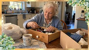 A woman in her 80s sitting at her kitchen table calmly sorting one small drawer with a cup of coffee beside her