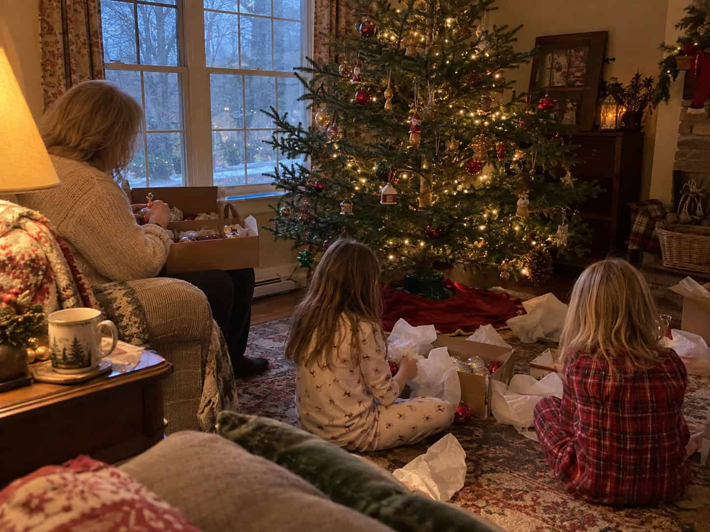 A woman in an armchair with an ornament box on her lap while two young girls unwrap ornaments on the rug beside a half-decorated Christmas tree.
