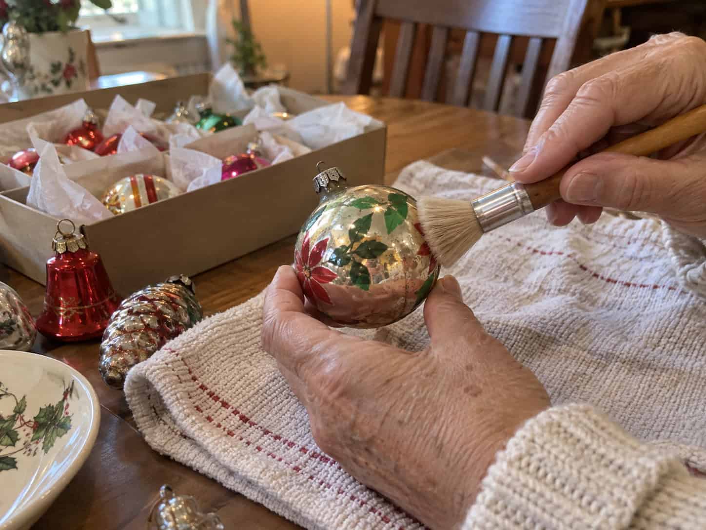 A woman's hand holding a vintage glass ornament by its body while lightly dusting it with a soft dry paintbrush.