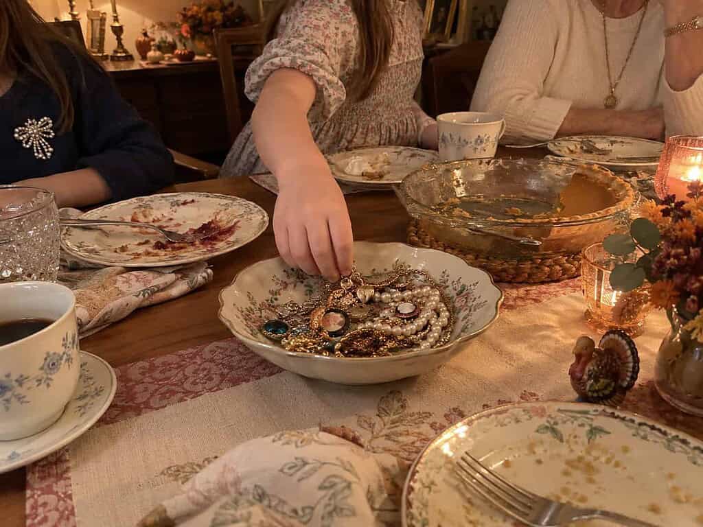 A child's hand reaching for a charm bracelet in a dish of costume jewelry on a dining table after Thanksgiving dinner, with a woman seated nearby.