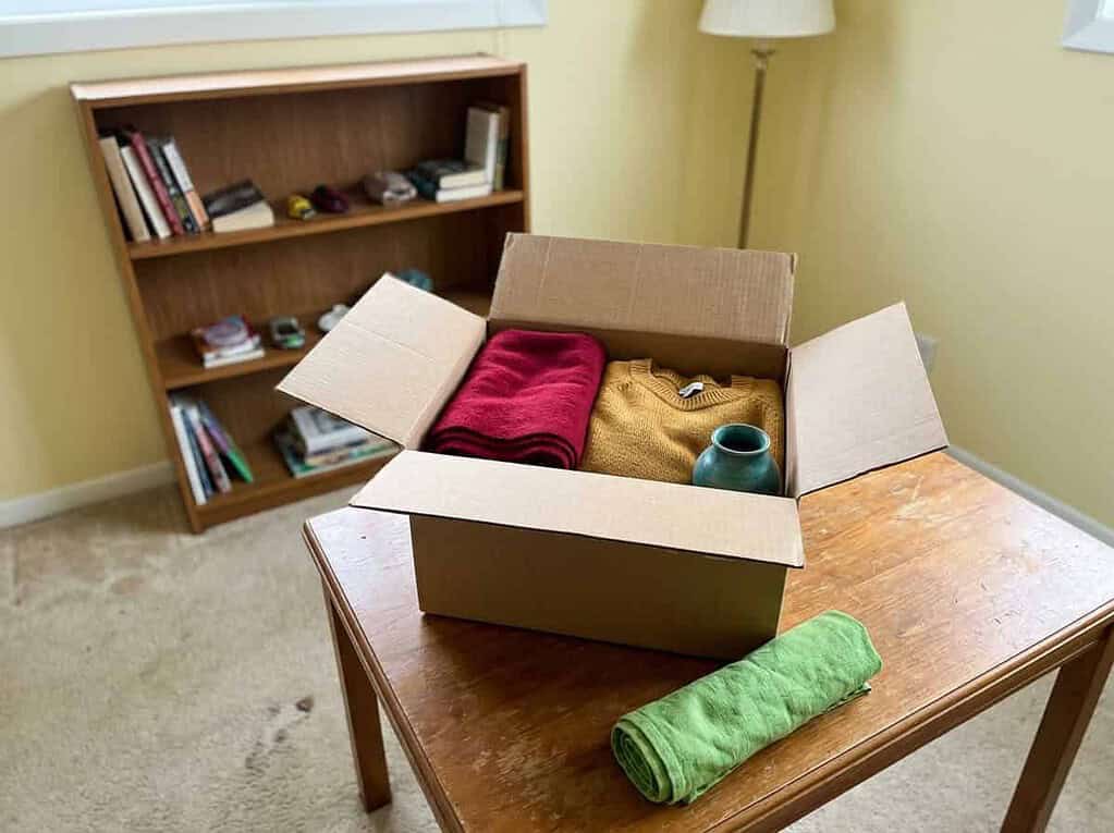 An open donation box with a few items inside next to a half-cleared shelf, gently loosening the grip on belongings