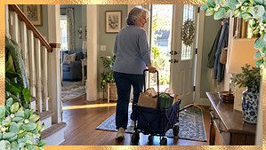 A woman in her 60s rolling a small cart of half-full donation bags toward her front door without lifting them
