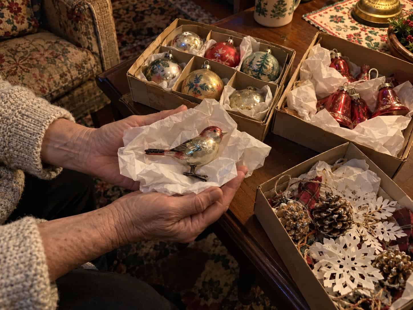 A woman's hands holding a small vintage glass bird ornament on white tissue, next to three boxes of old family ornaments.