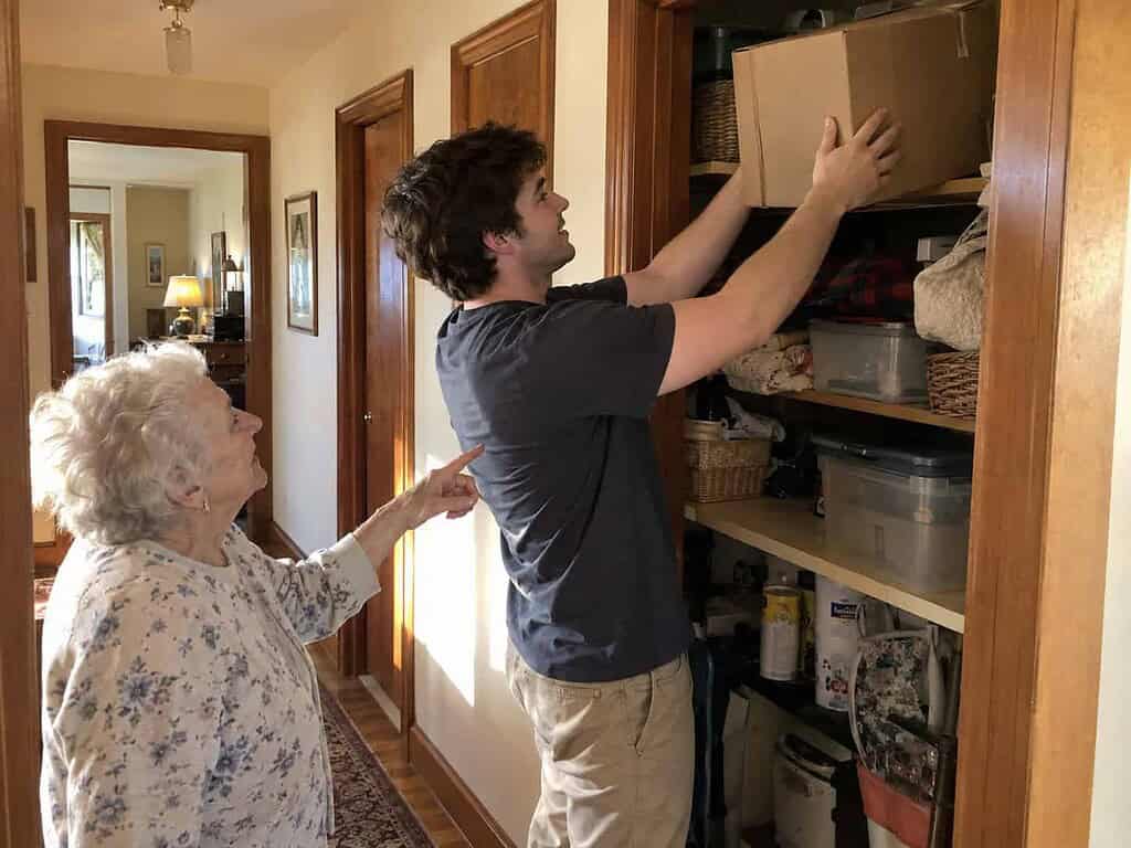 A young man lifting a box down from a high closet shelf while a woman in her 80s stands safely nearby