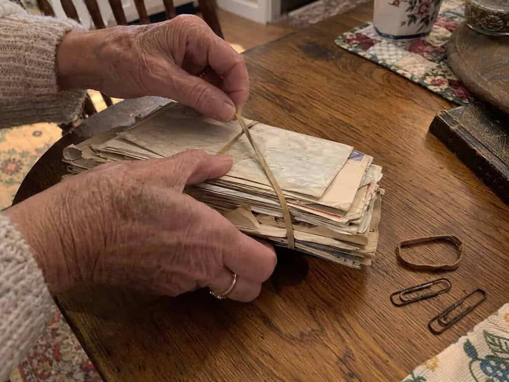 An older woman's hands removing a dried rubber band and rusty paper clips from a stack of old letters