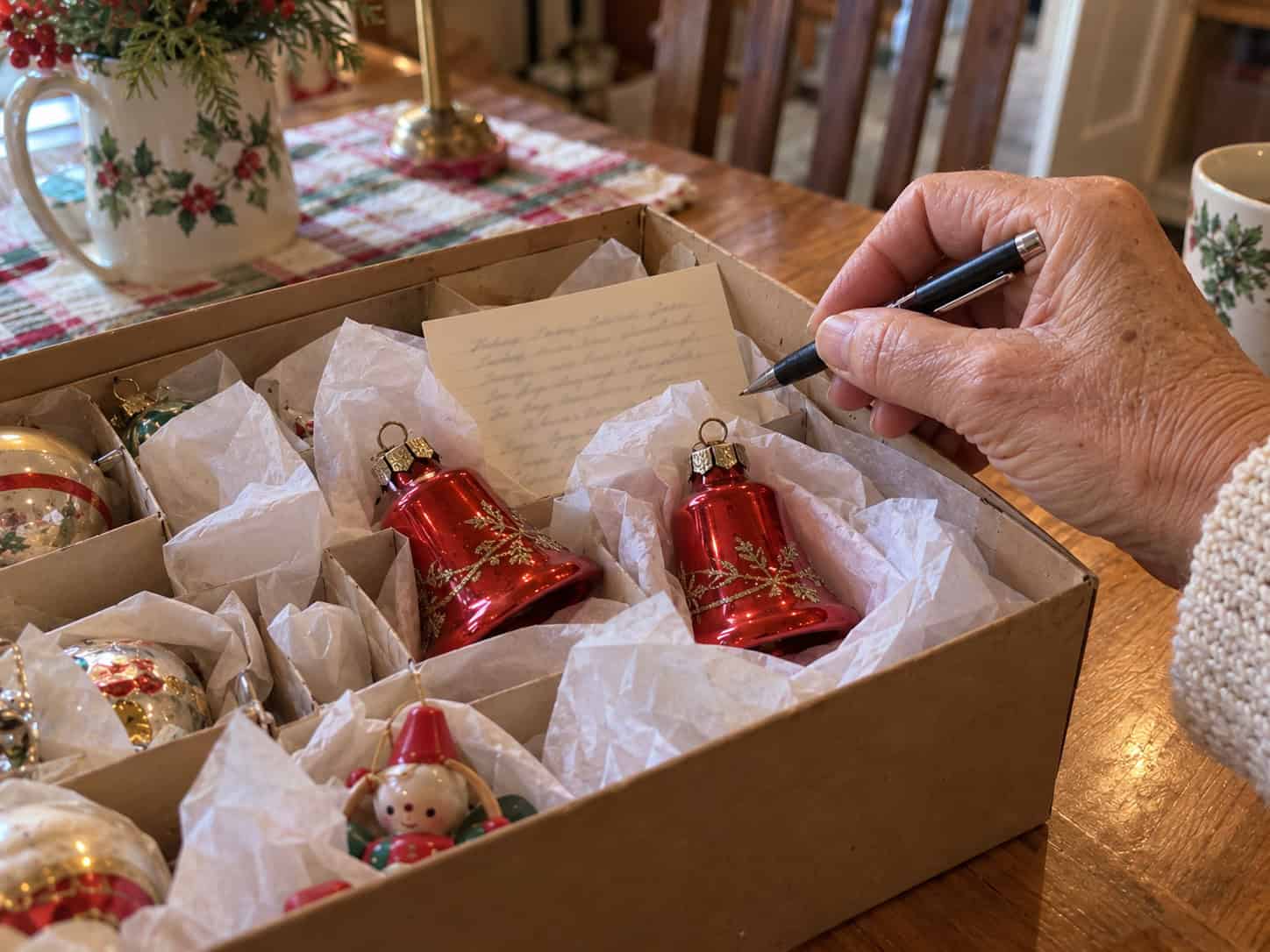 A handwritten note card tucked inside a box of red glass bell ornaments, with a woman's hand holding a pen beside it.