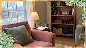 A warm shared living room with a calm armchair corner and a husband's tidy shelf of kept things nearby