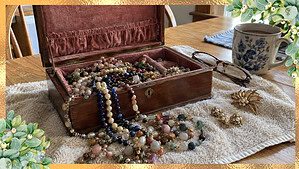 Old wooden jewelry box open on a kitchen table with tangled necklaces, clip-on earrings, a gold brooch, and reading glasses on a towel.