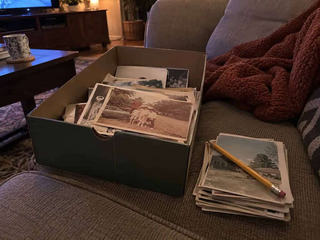 One open shoebox of old photos on a couch with a small neat stack sorted beside it and a pencil on top, in cozy evening light