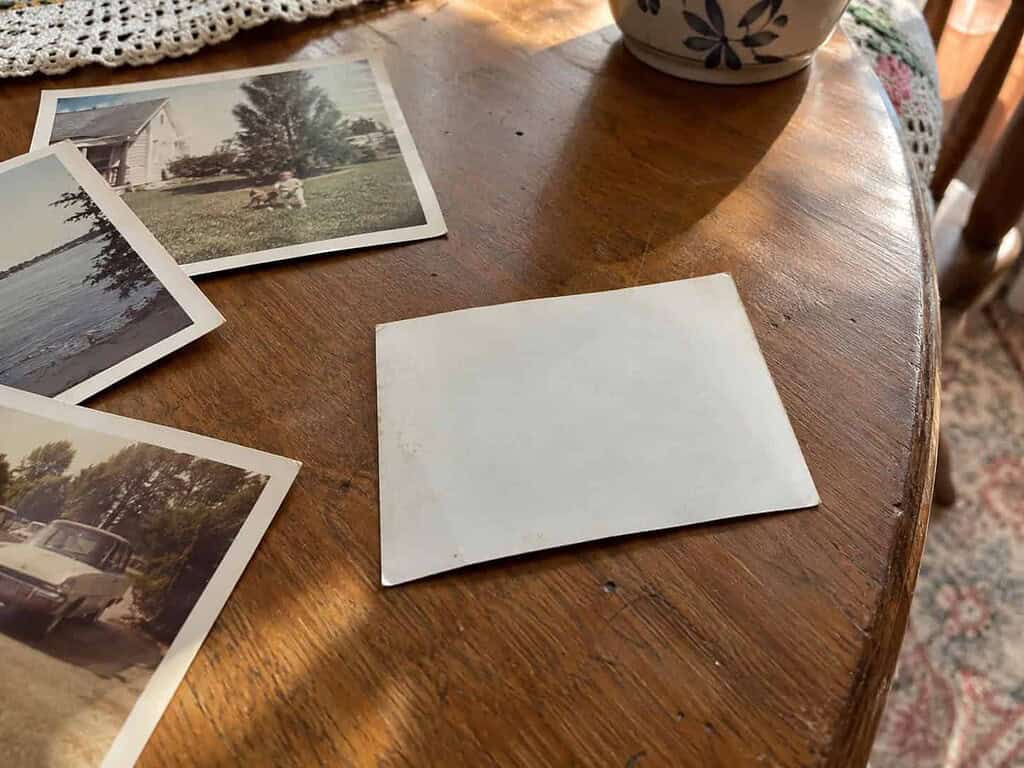 An old family photo turned face-down showing a blank, unlabeled back on a wooden table with scattered loose prints