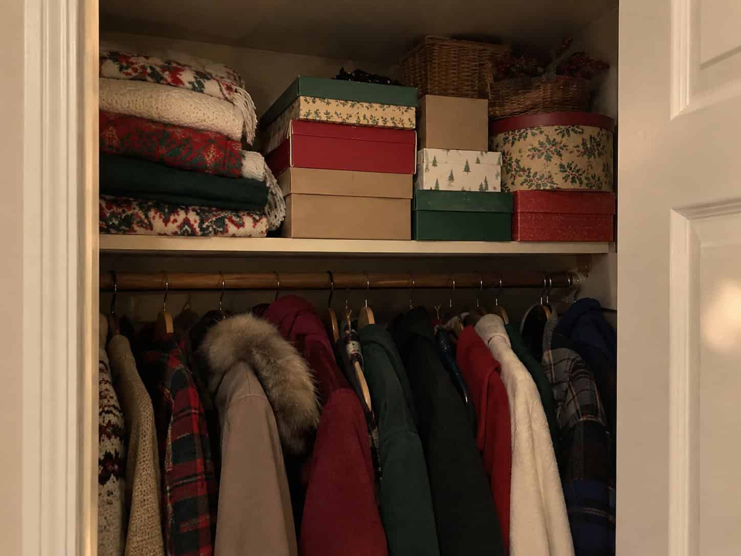 Cardboard ornament boxes stacked on a hall closet shelf next to folded blankets, with coats hanging below.