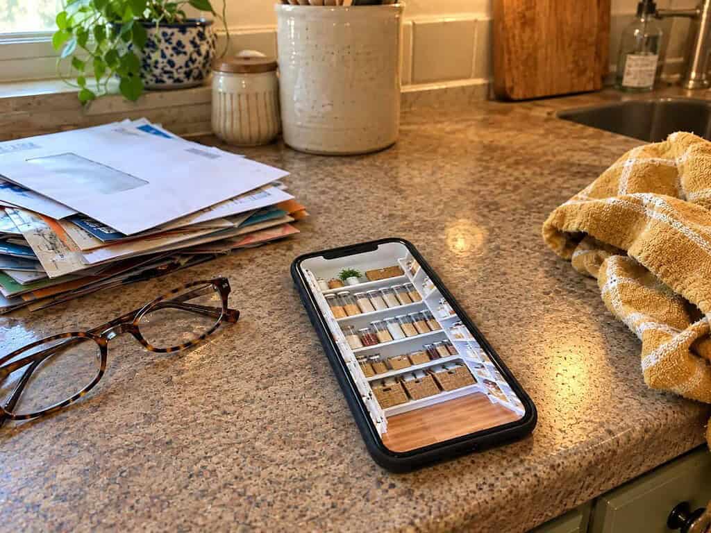 A phone showing a spotless organized pantry propped on a real counter cluttered with mail, glasses, and a dish towel