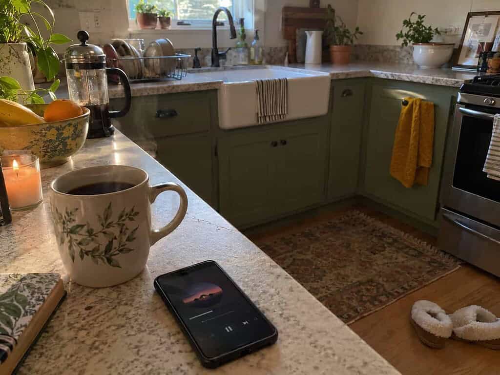 A kitchen counter set up for a slow reset morning with a steaming coffee mug, a phone playing music, and comfy slippers