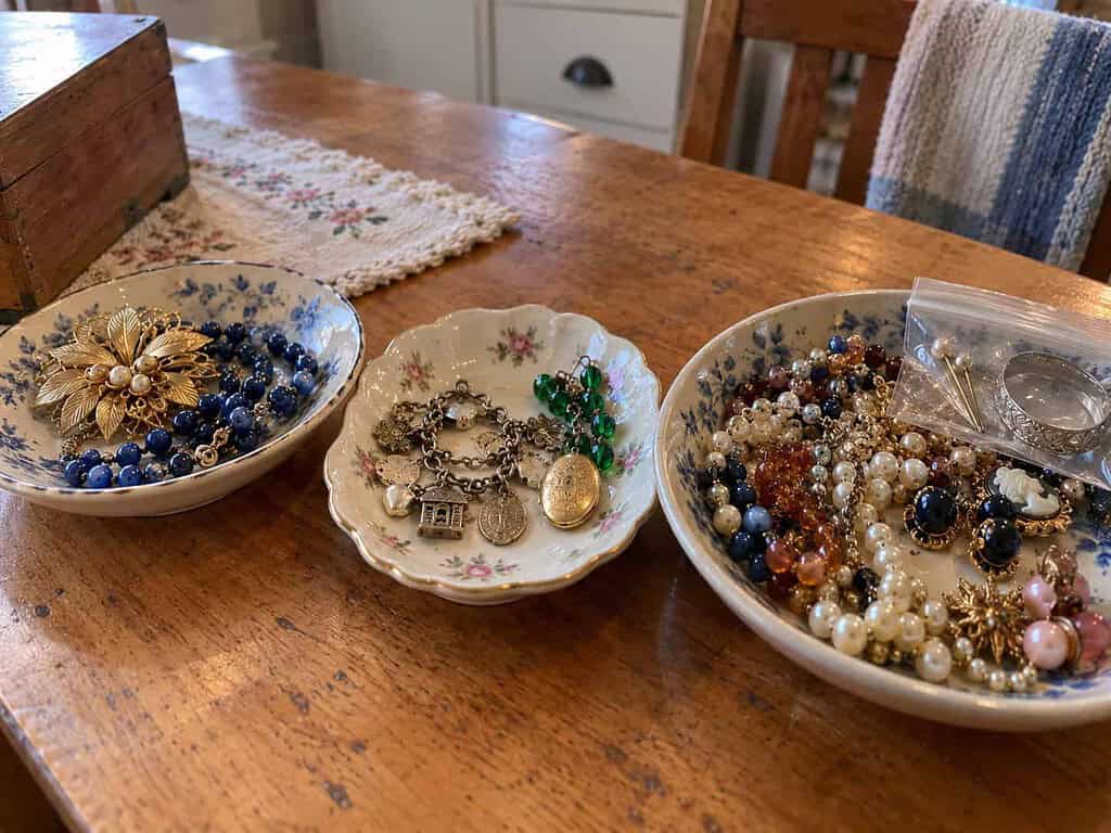 Three shallow dishes on a kitchen table holding sorted costume jewelry, with a small zip bag of marked pieces in one dish.