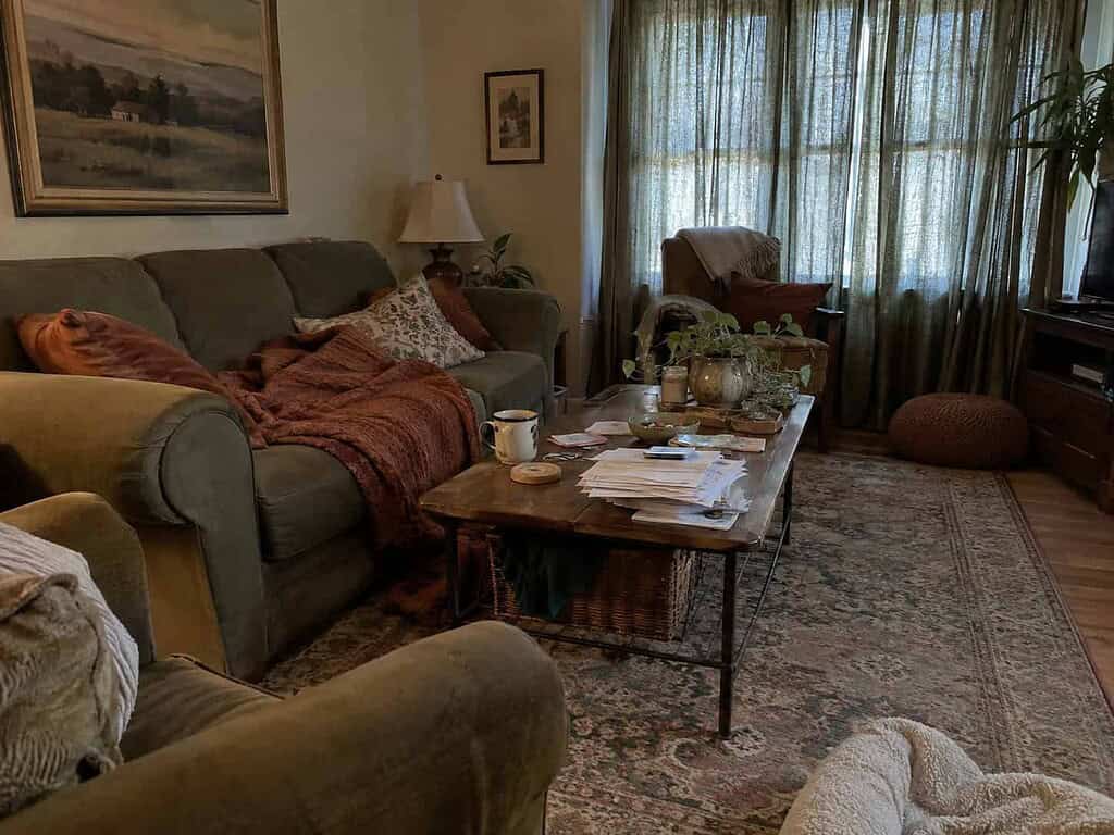 A tired-looking living room before a reset, with a cluttered coffee table, a wadded blanket, and drawn curtains