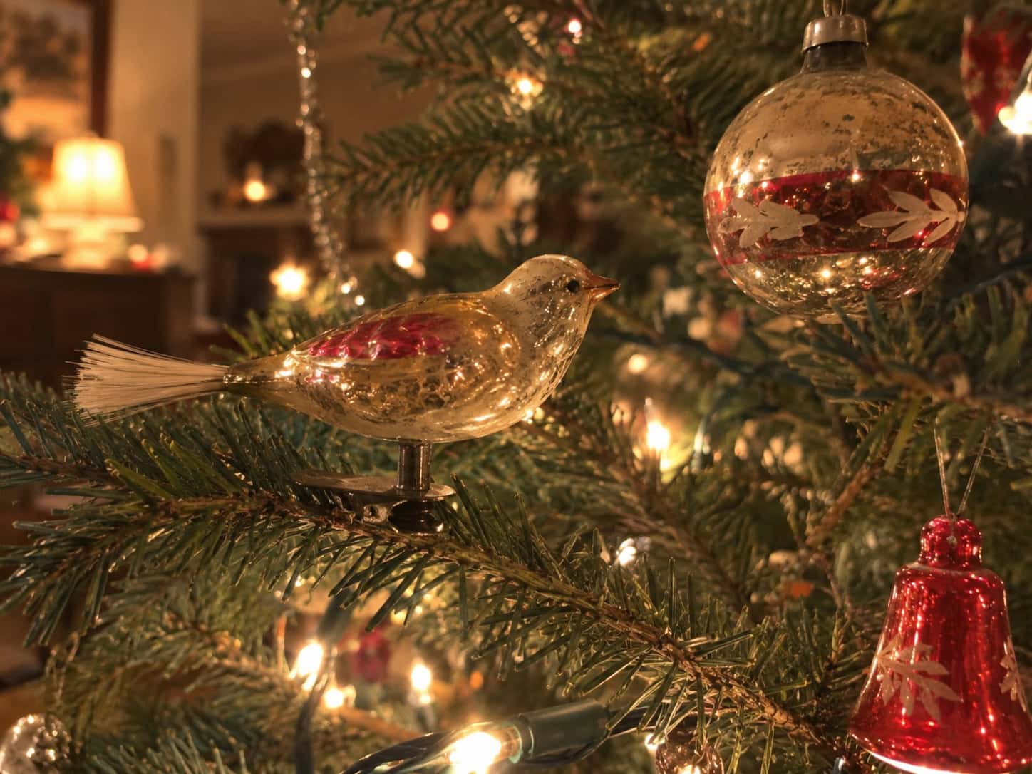 A small vintage glass bird ornament clipped to a Christmas tree branch, with soft string lights glowing behind it.