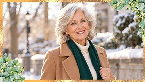 Woman in her late seventies with silver hair wearing a camel coat over a cream knit with a jewel-tone scarf on a bright winter day.