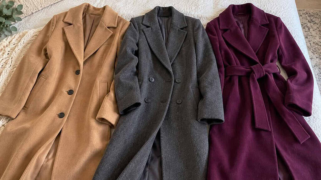A camel wool coat, a charcoal tailored coat, and a deep plum belted coat laid together on a pale neutral surface.