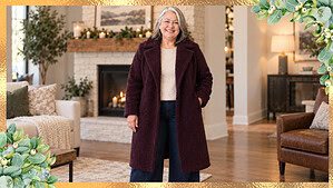 Woman in her fifties with silver hair wearing a cream knit sweater, dark wide-leg jeans, and an open deep-plum winter coat with cognac boots, standing in a cozy living room.
