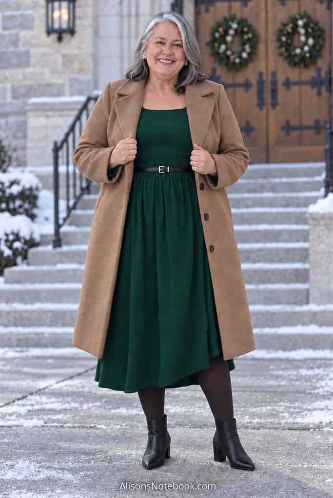 Woman in her fifties with silver hair wearing an emerald green long-sleeve midi dress with a belted waist, espresso tights, and an open camel coat with black pointed ankle boots, standing at a church entrance.