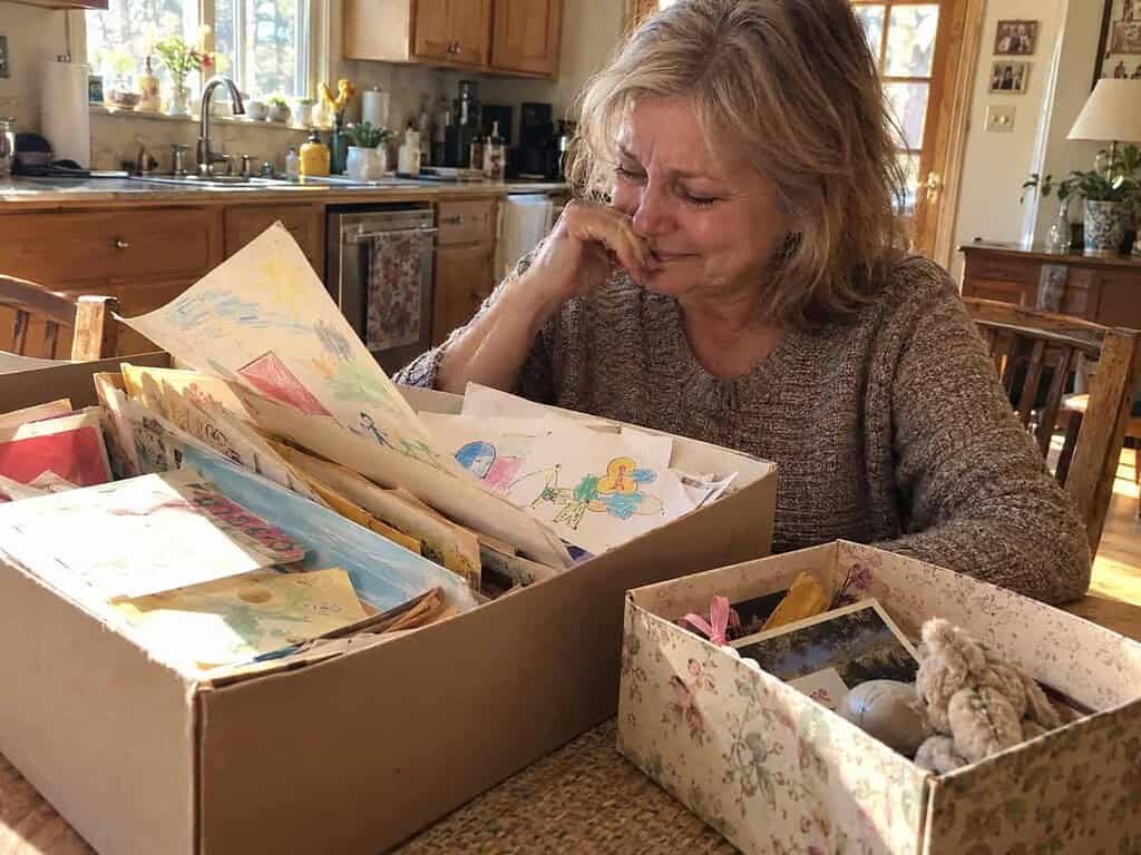 A woman in her 60s smiling with teary eyes as she sorts her grown children's old school drawings at the kitchen table