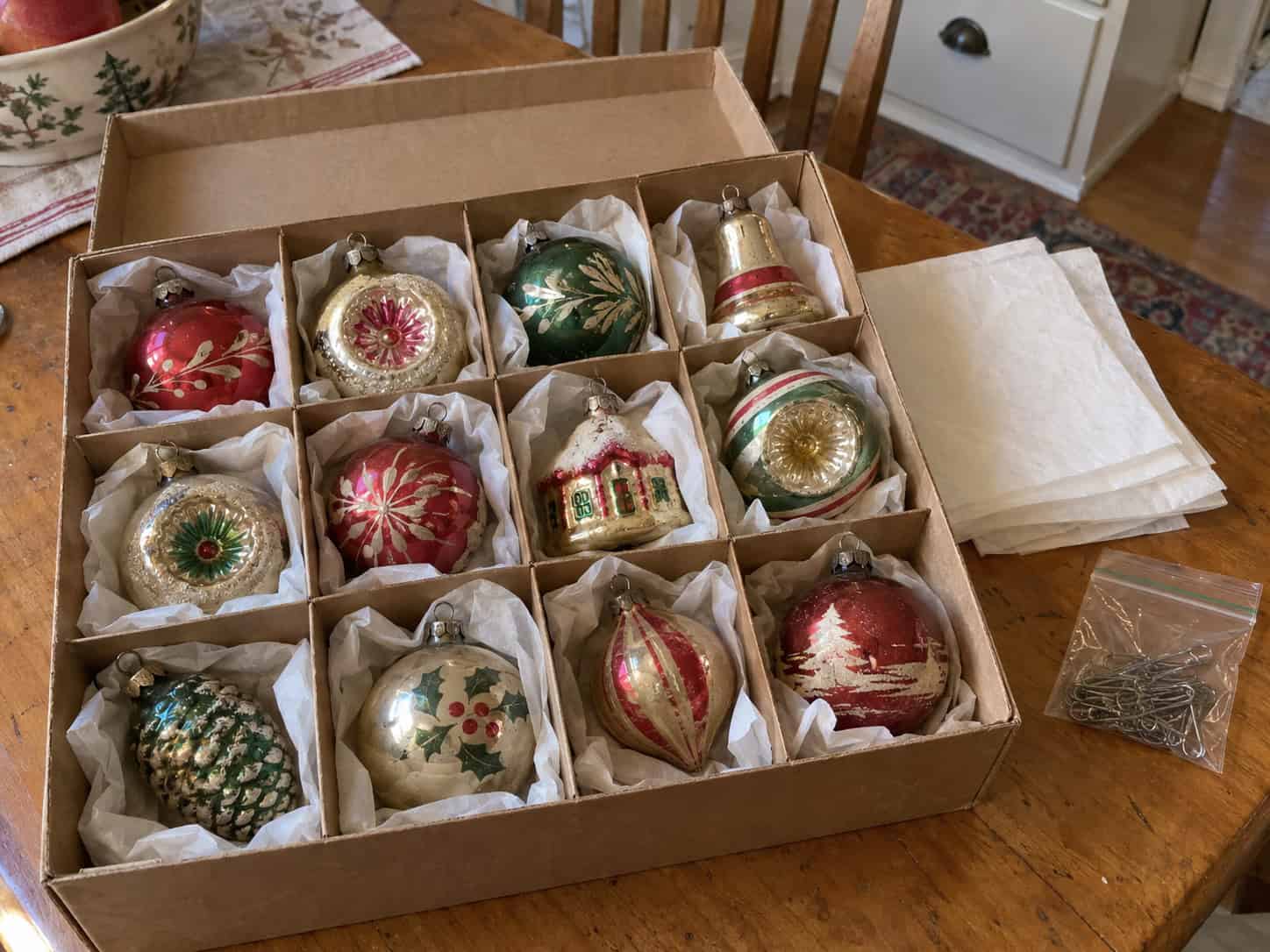 A divided cardboard box holding old glass ornaments loosely wrapped in white tissue, with a small bag of metal hooks beside it.