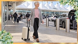 Woman over 60 in a gray cardigan, red striped tee and loafers pulling a carry-on suitcase outside the airport
