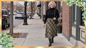 Ankle boots outfits over 60: silver-haired woman in a black turtleneck, olive plaid midi skirt, tights, and black block-heel ankle boots on a town sidewalk