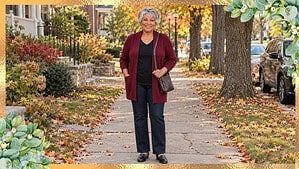 Smiling woman in her 60s with an apple shape in a long wine-red cardigan, black tee and dark jeans on a fall sidewalk