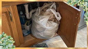 A large plastic bag stuffed full of grocery bags, bulging out of an open cabinet under a kitchen sink
