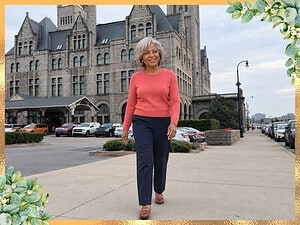 Smiling woman with a gray bob in a coral sweater and navy trousers walking past a historic stone train station