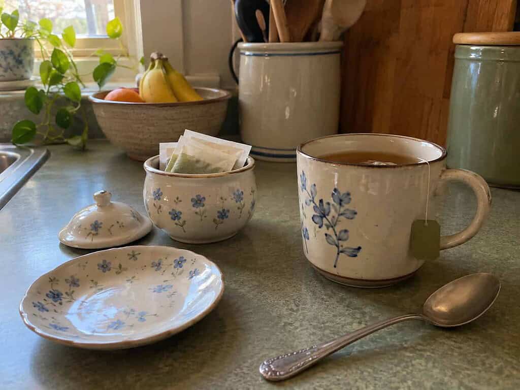 A vintage blue-flowered sugar bowl holding tea bags on a kitchen counter, beside a mug of tea and a matching small plate.