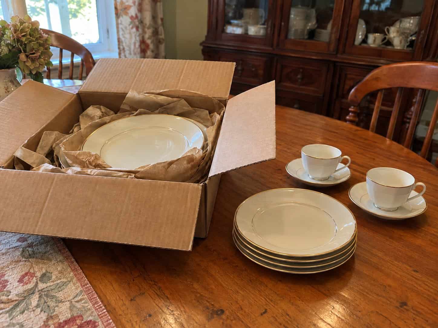 An open box of gold-rimmed china in packing paper on a dining table beside a stack of plates and two teacups