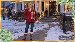 Smiling woman over 60 in a red peacoat, plaid scarf, dark jeans and cuffed brown boots outside a house with Christmas lights