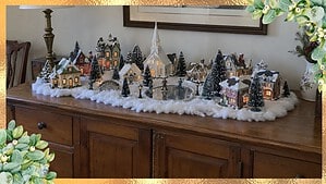 A lighted ceramic Christmas village with a steepled church, a mirror skating pond and bottle-brush trees on cotton snow on a dining room buffet
