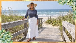 Smiling woman over 60 in a navy striped boat-neck top, white linen maxi skirt, straw hat and brown sandals on a beach walkway