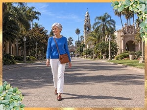 Smiling woman with a silver pixie cut wearing a cognac crossbody bag, blue sweater and white jeans on a sunny park promenade