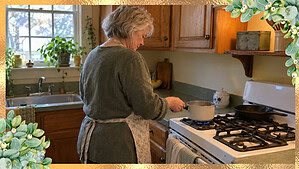 A woman in her 60s with silver hair holds a small saucepan at a white gas stove, with a cast-iron skillet on the back burner
