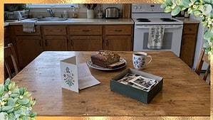 Birthday card with a handwritten list tucked inside, banana bread, coffee and a shoebox of old photos on a kitchen table