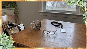 A cup of tea, reading glasses and a work badge on a lanyard on a clear kitchen table, with a work tote bag on the chair