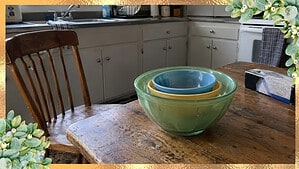 A stack of old colorful mixing bowls on a wooden kitchen table beside a box of trash bags, with a chair pulled out
