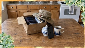 Old fishing hat with lures resting on an open box from the closet beside a phone and coffee on a kitchen table