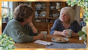 Two women in their 60s sharing coffee at a kitchen table, with a china cabinet of pretty dishes behind them.
